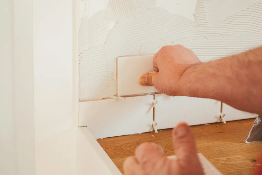 Hands setting a white tile on a bathroom wall
