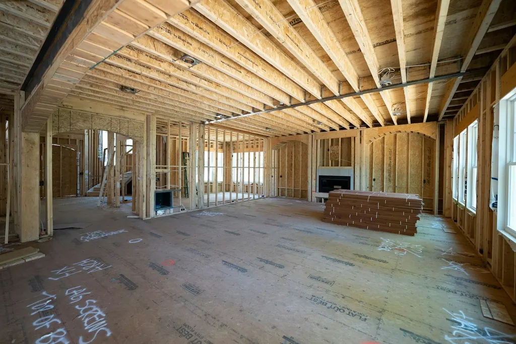 A wood-framed interior with floor joists overhead and stacked lumber, before drywall